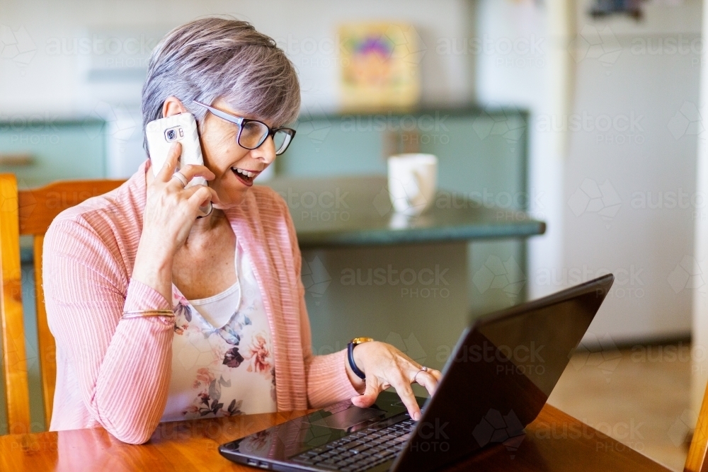 Mature woman on laptop at home using mobile phone to call for tech support - Australian Stock Image