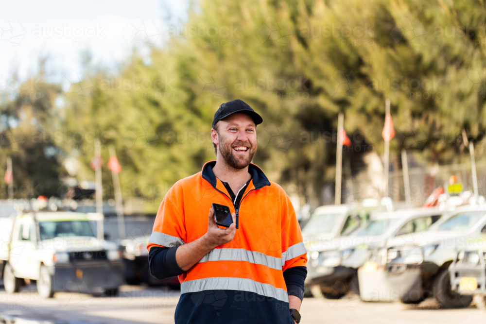 Mechanic workman in high-vis holding a mobile device on a phone call outside industrial workshop - Australian Stock Image