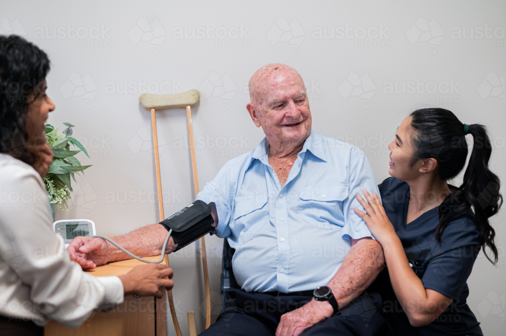 Medical practitioner taking elderly mans blood pressure while female caregiver crouched beside him - Australian Stock Image
