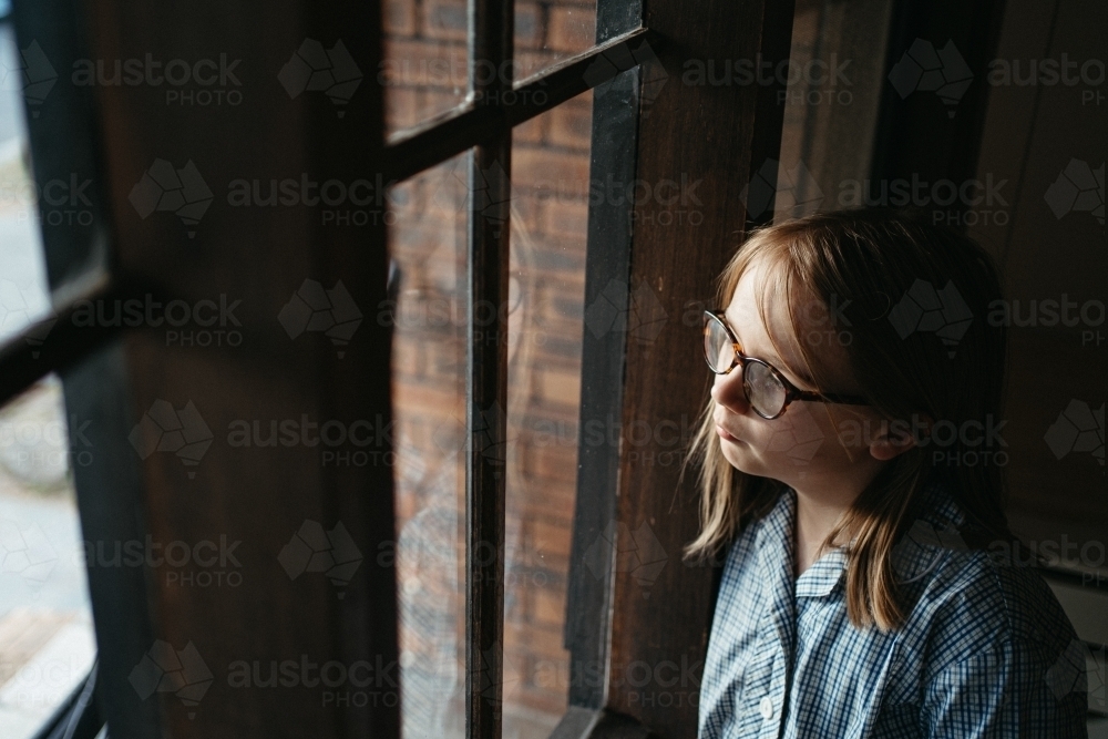 Melancholic primary aged girl in a school uniform looks out a window - Australian Stock Image