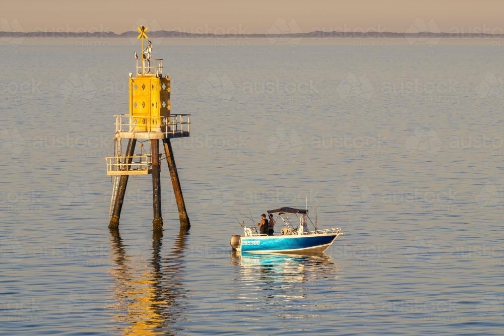 Men fishing from a small boat on calm water near a channel marker - Australian Stock Image