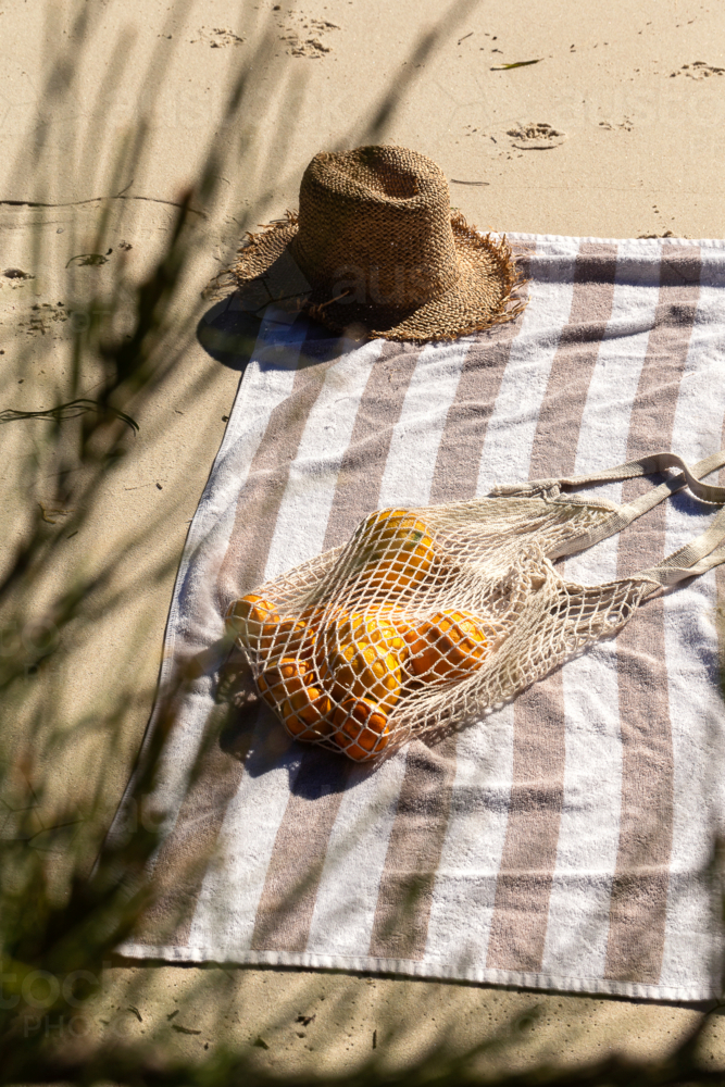 Mesh bag of fruits and straw hat on top of striped beach towel on the sand - Australian Stock Image