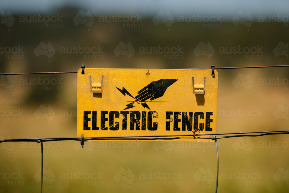 Metal fence with yellow 'Electric Fence' sign in rural landscape under clear blue sky - Australian Stock Image