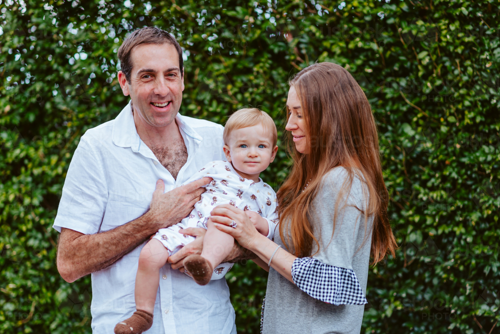 mid aged man and woman holding up a young boy with blond hair in front of a green background - Australian Stock Image
