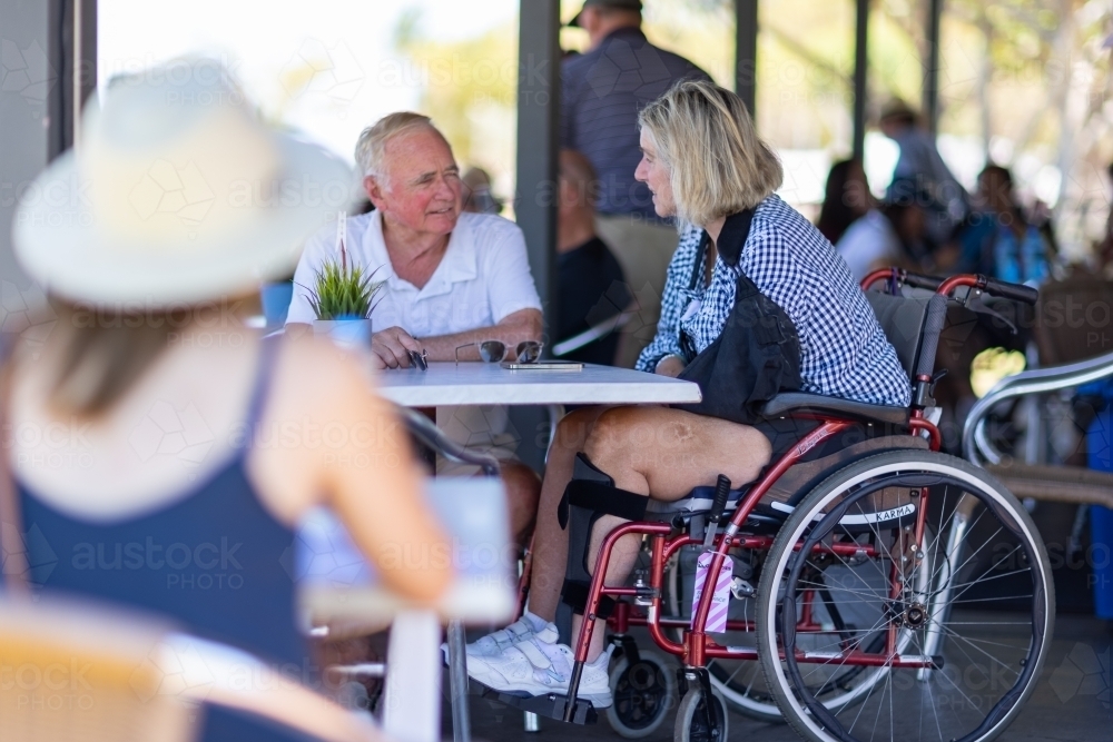 middle aged couple sitting at cafe table with woman in wheelchair - Australian Stock Image