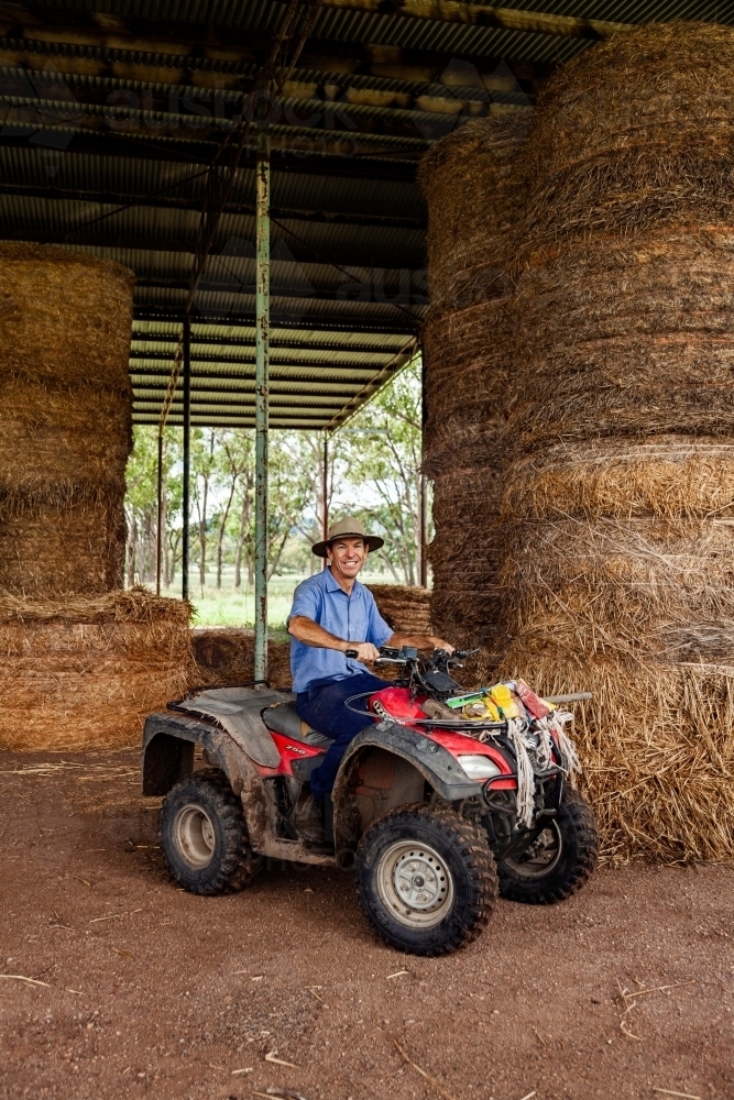 Middle aged farmer on quad bike at farm hay shed collecting feed for cattle - Australian Stock Image