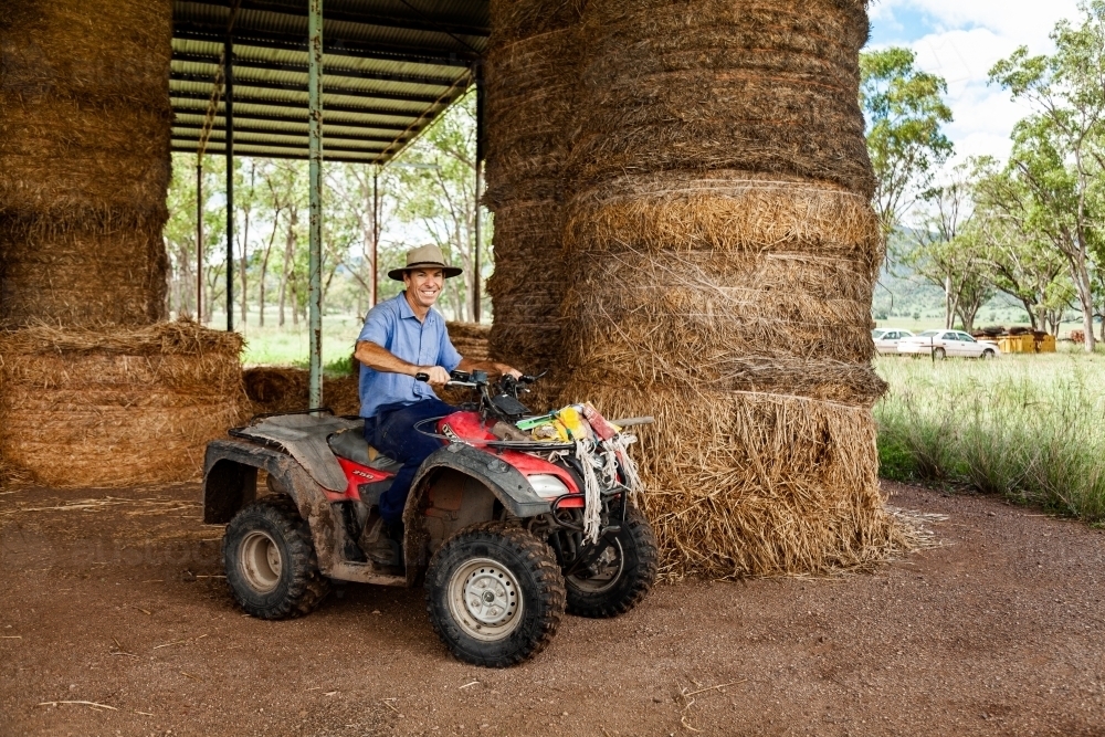 Middle aged farmer on quad bike at farm hay shed collecting feed for cattle - Australian Stock Image