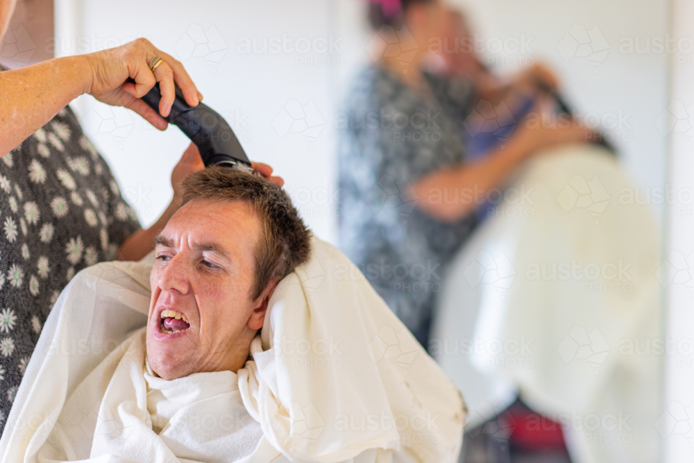 Middle aged woman carer cutting hair on head of young man with disability - Australian Stock Image