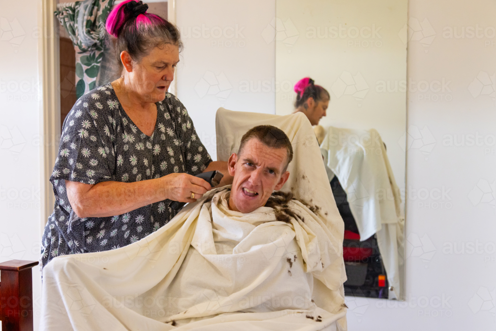 Middle aged woman carer cutting hair on head of young man with disability - Australian Stock Image