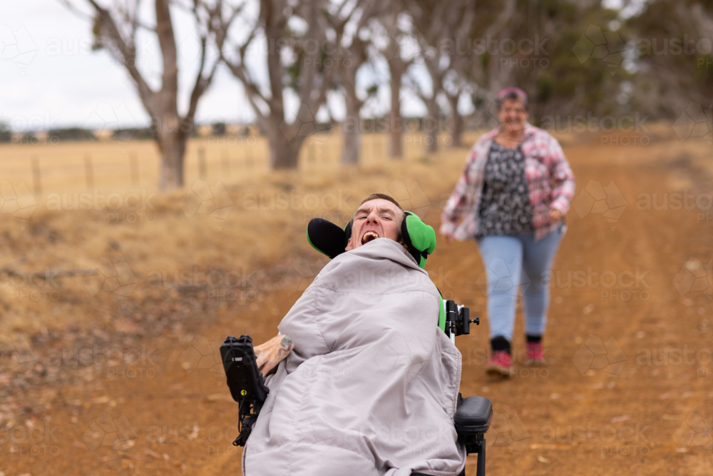 Middle-aged woman running after her son with disability on wheelchair on dirt road - Australian Stock Image