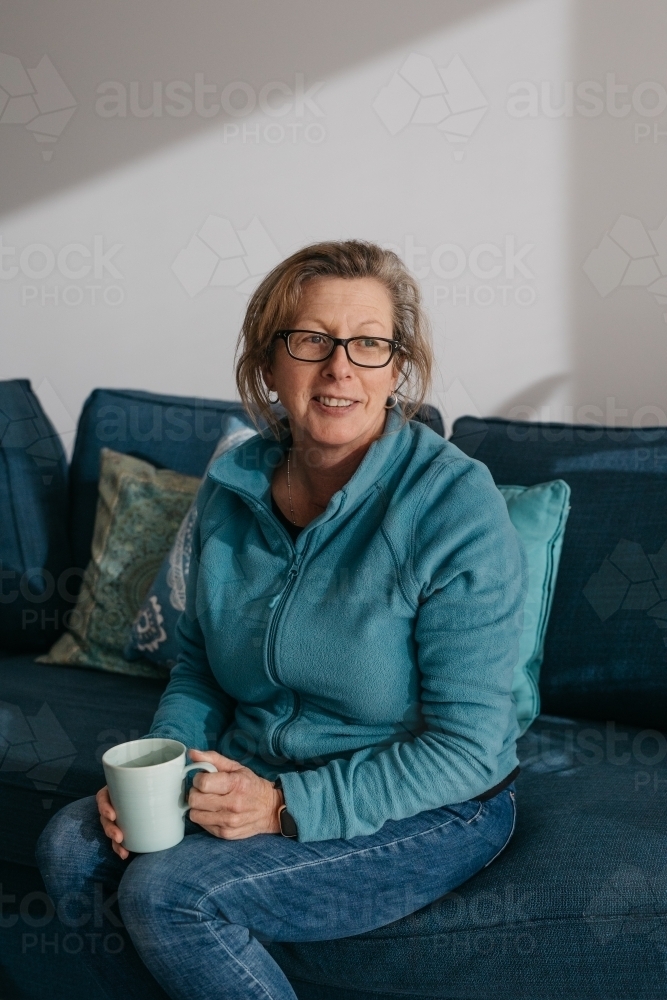 Middle aged woman sitting on couch, drinking hot beverage - Australian Stock Image