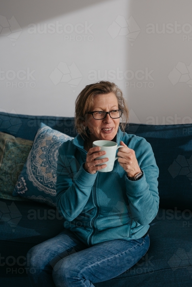 Middle aged woman sitting on couch, drinking hot beverage - Australian Stock Image