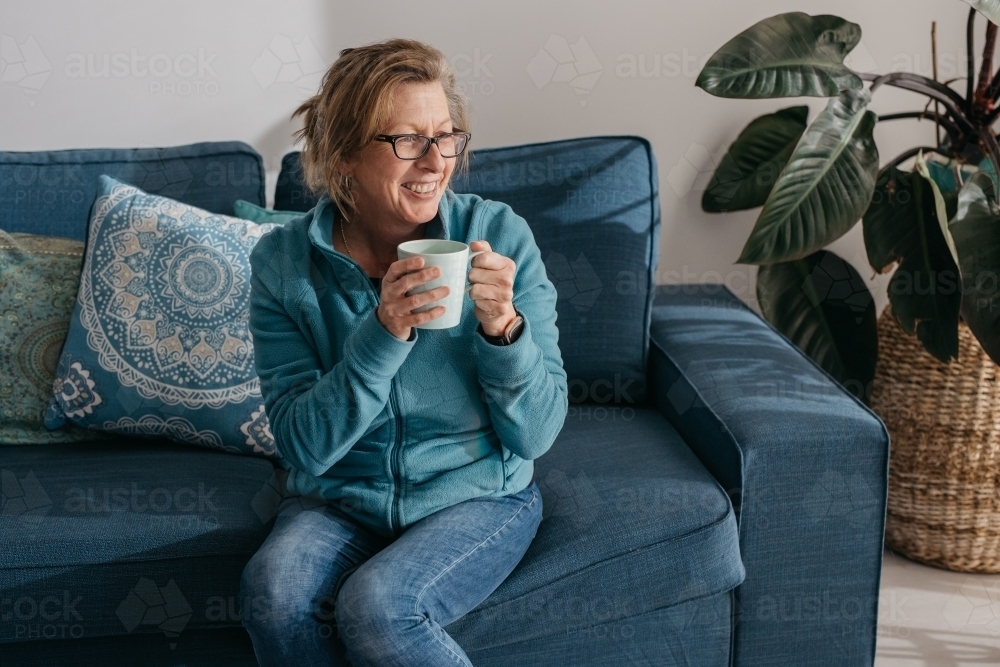Middle aged woman sitting on couch, drinking hot beverage - Australian Stock Image