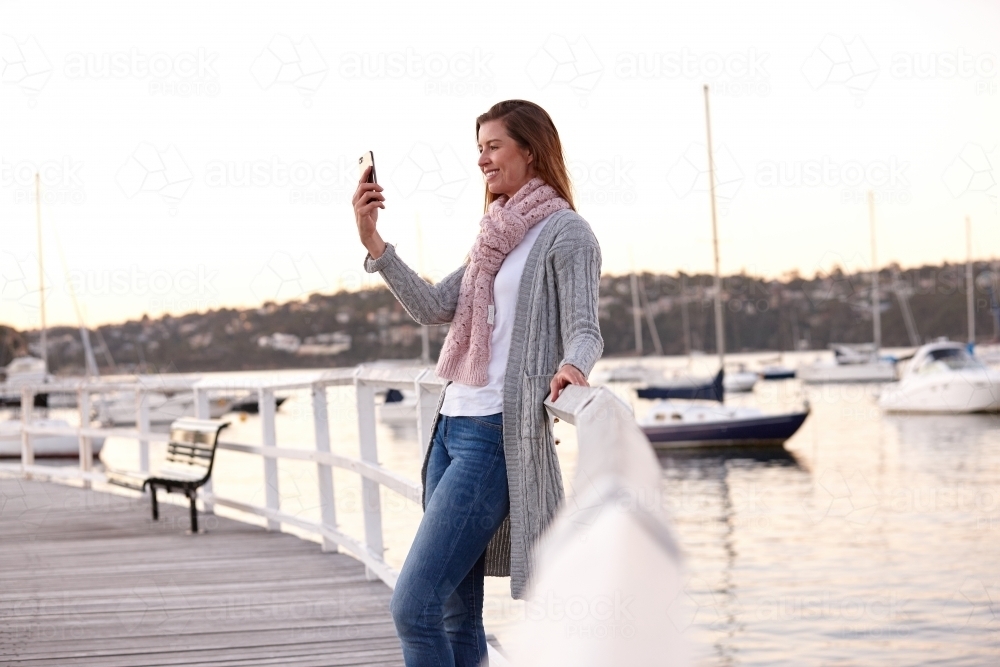 Middle aged woman using mobile phone on wharf to take selfie - Australian Stock Image