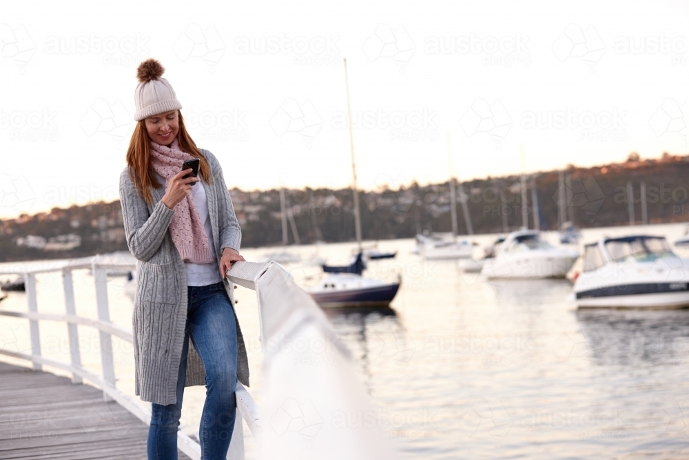 Middle aged woman using mobile phone on wharf wearing beanie - Australian Stock Image