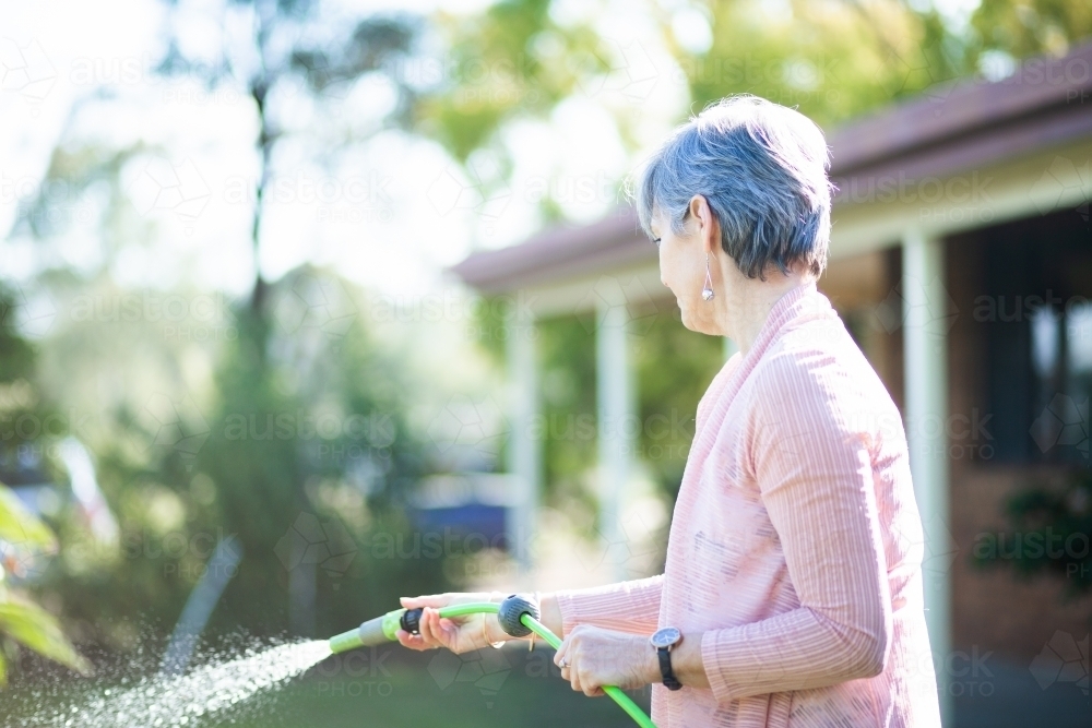 Middle aged woman watering her garden beside her house - Australian Stock Image