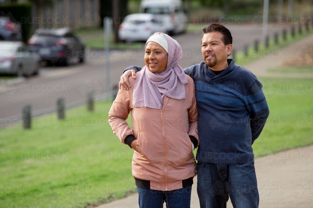 middle aged woman wearing pink hijab and middle aged man wearing blue sweater putting his arm on her - Australian Stock Image