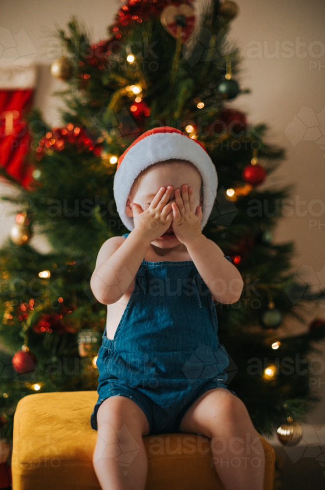 Mischievous blonde girl plays peekaboo wearing a Santa hat by a Christmas tree - Australian Stock Image