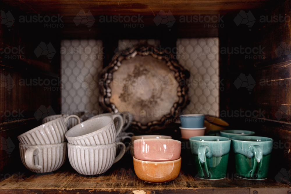 Mix of ceramic cups, bowls, and vintage dishware neatly arranged wooden cabinet - Australian Stock Image