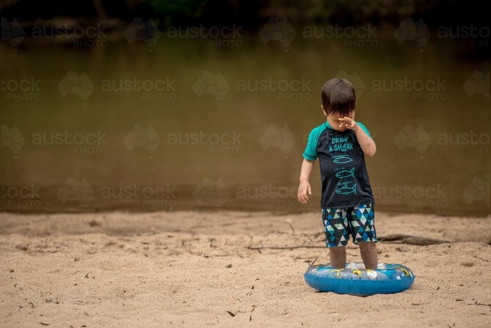 Mixed race 3 year old boy plays on a river bank with an inflatable swimming tube - Australian Stock Image