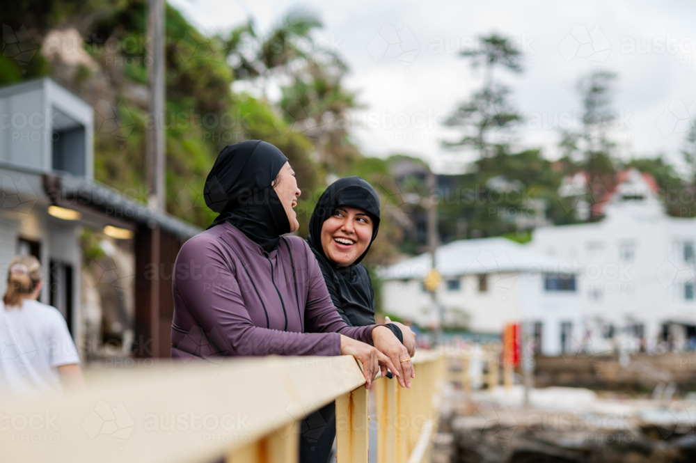 Mixed race women standing and leaning on the railings along coastal path while chatting - Australian Stock Image