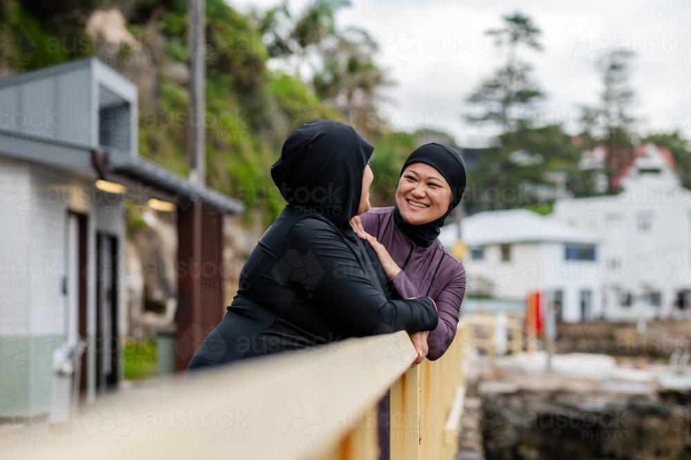 Mixed race women standing and leaning on the railings along coastal path while chatting - Australian Stock Image
