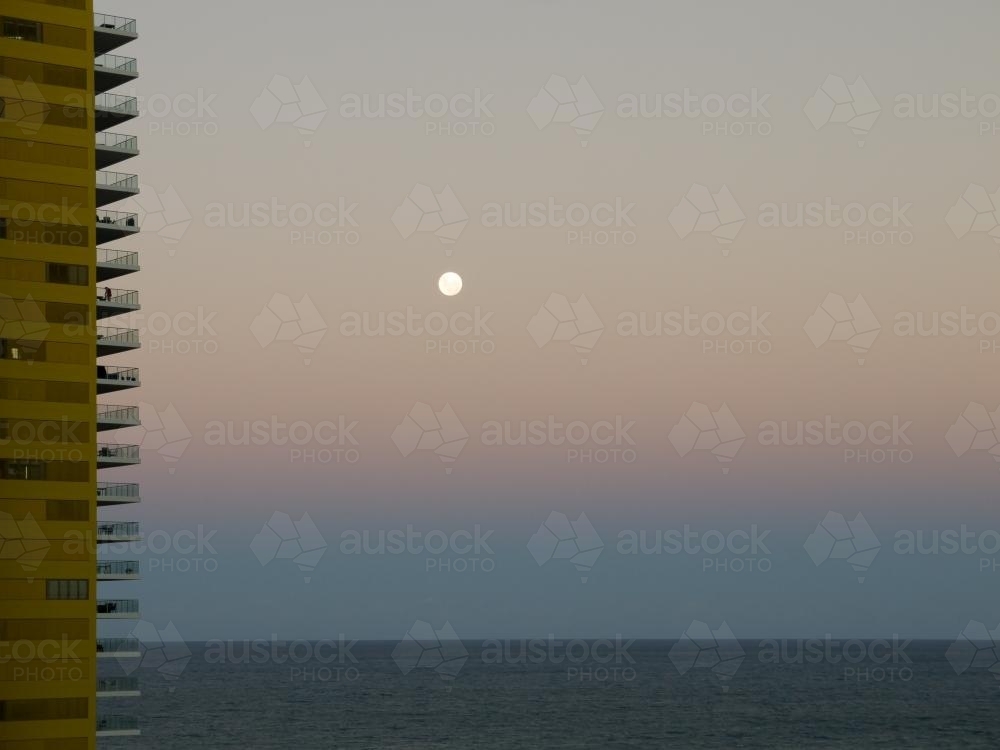 Moon rising over the sea at dusk with tall building on left - Australian Stock Image