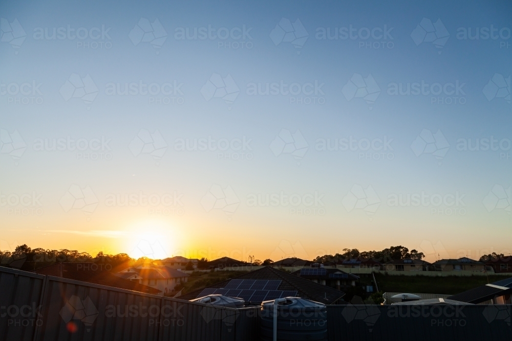 Morning sun rising in clear sky over backyard fence and water tank with houses and rooves - Australian Stock Image