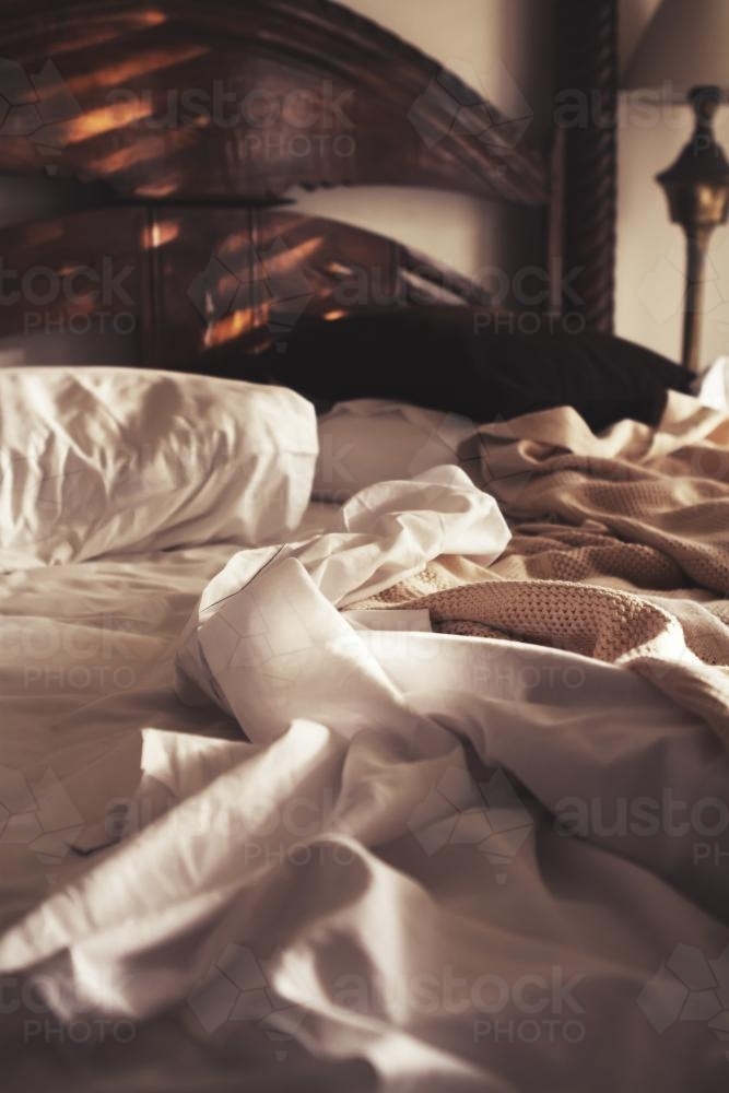Morning sunlight on an unmade luxury bed of white sheets and beige blanket - Australian Stock Image