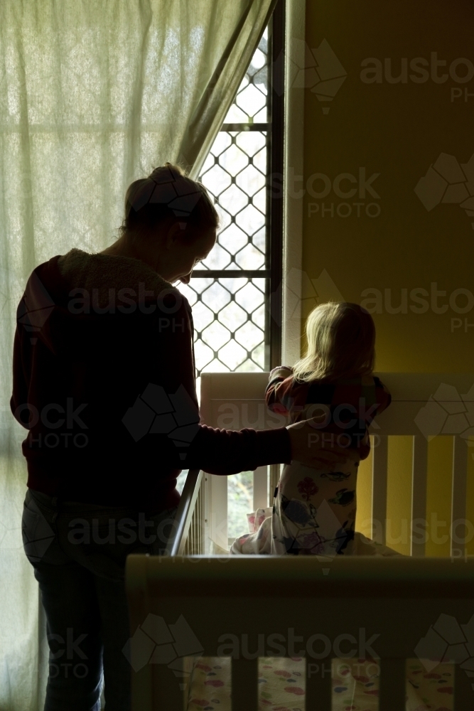 Mother and baby daughter in the bedroom as she stands and looks out window. - Australian Stock Image