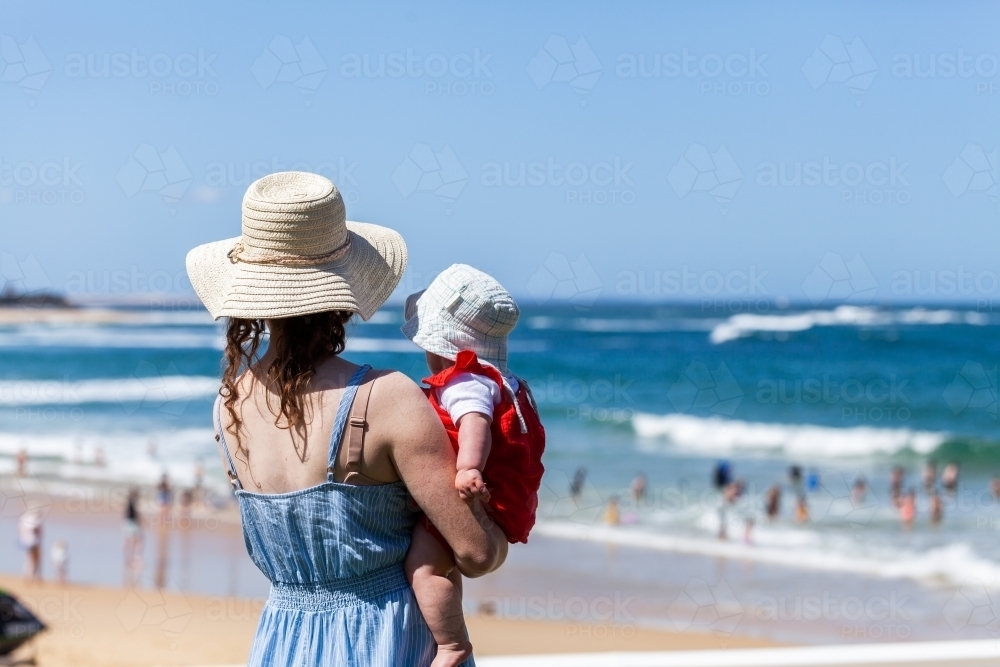Mother and baby wearing a hat looking out over busy beach in summer - Australian Stock Image