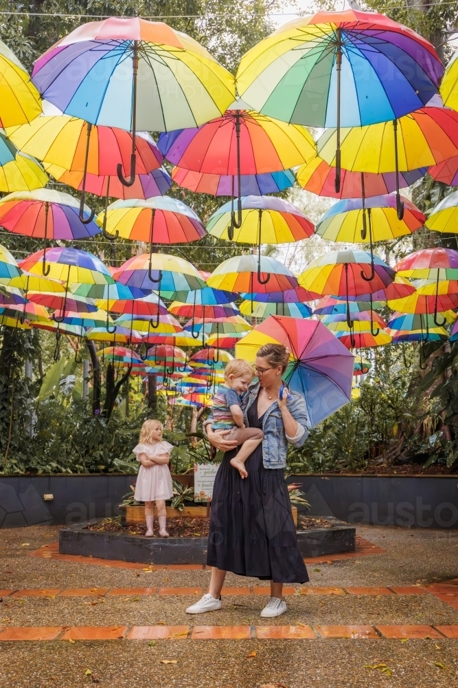 Mother and children under bright rainbow umbrella art installation in the rain - Australian Stock Image