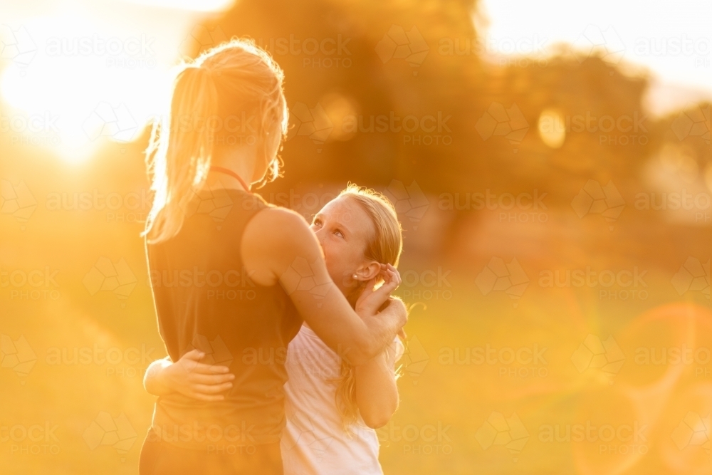 mother and daughter backlit and bathed in golden light with sun flare embracing outdoors - Australian Stock Image