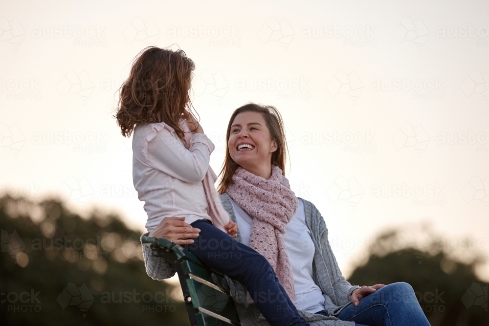 Mother and daughter sitting on park bench at park on sunset - Australian Stock Image