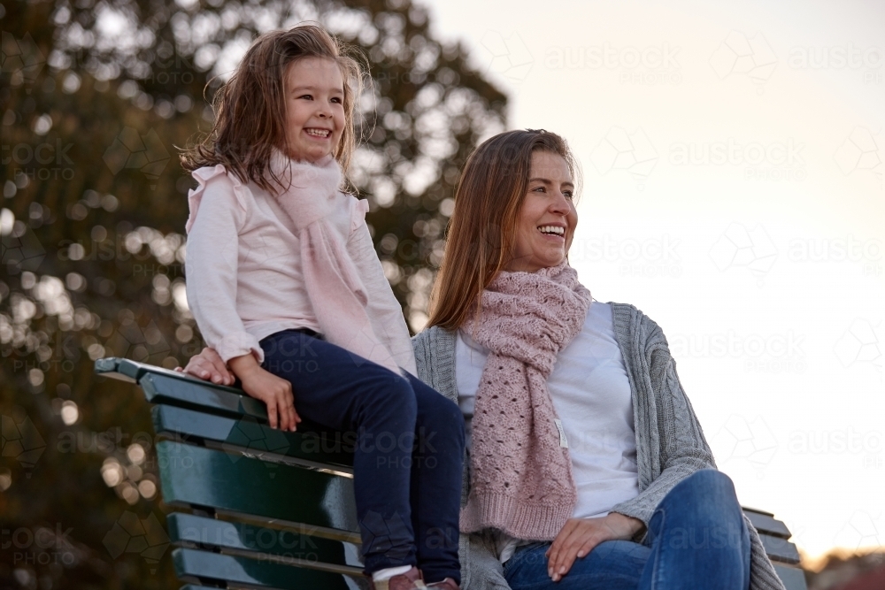 Mother and daughter sitting on park bench at park on sunset - Australian Stock Image