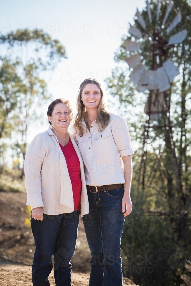 Mother and daughter standing smiling with windmill in background on farm in drought - Australian Stock Image