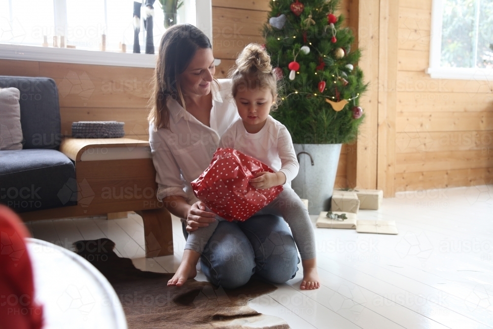 Mother and daughter unwrapping present with Christmas tree in background - Australian Stock Image