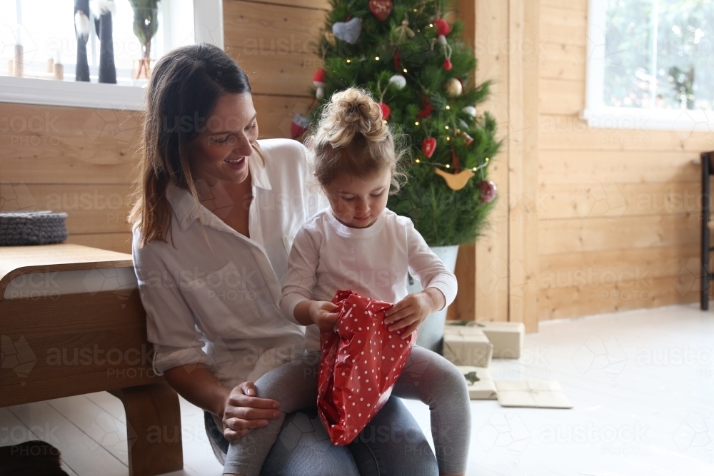 Mother and daughter unwrapping present with Christmas tree in background - Australian Stock Image
