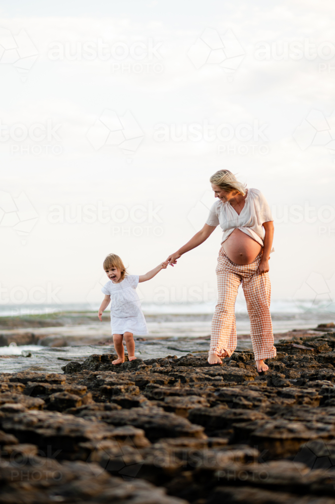 Mother and daughter walking along rocky coastline at sunset, expectant mother smiling joyfully - Australian Stock Image
