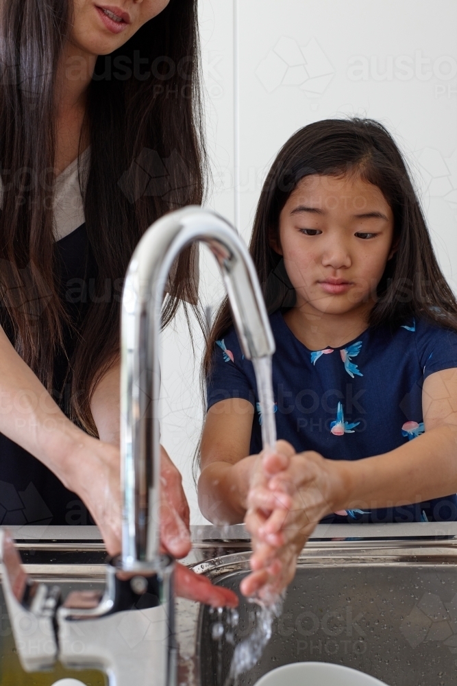 Mother and daughter washing hands together in kitchen sink - Australian Stock Image