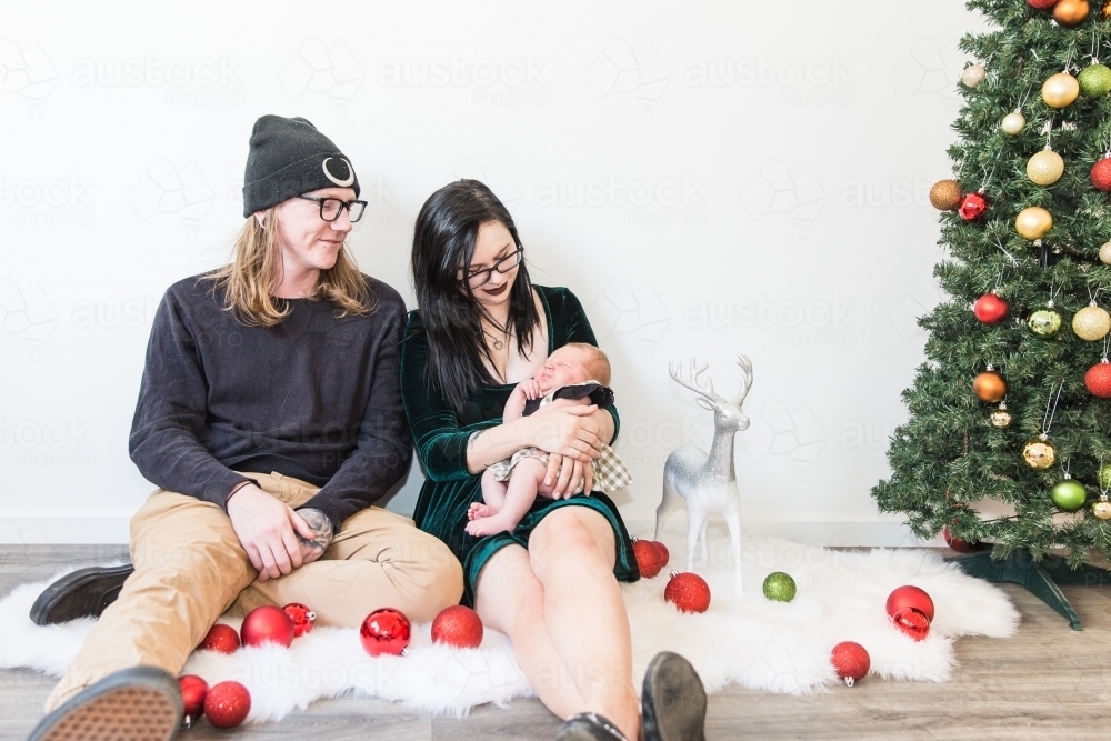 Mother and father smiling at baby in arms at Christmas with tree and decorations - Australian Stock Image