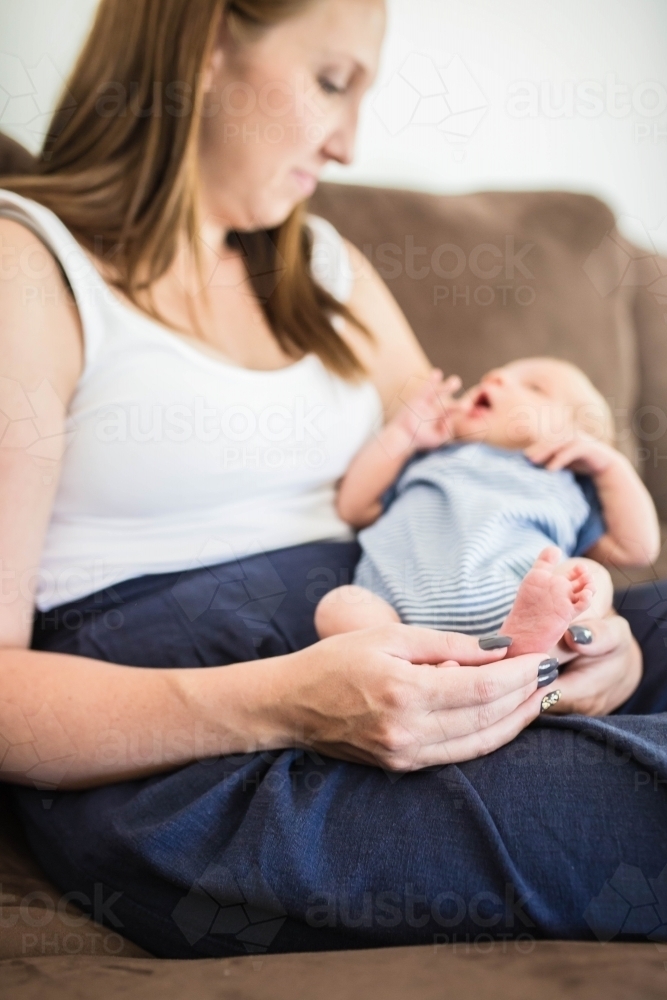 Mother and newborn baby boy sitting on lounge with mum holding son's feet - Australian Stock Image