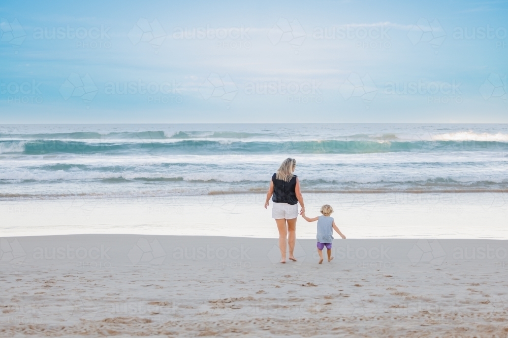 Mother and son holding hands walking towards water on the beach - Australian Stock Image