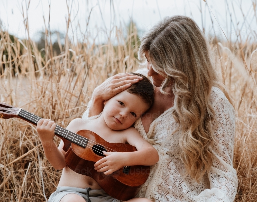 Mother and son sitting close together outside in field with small guitar - Australian Stock Image