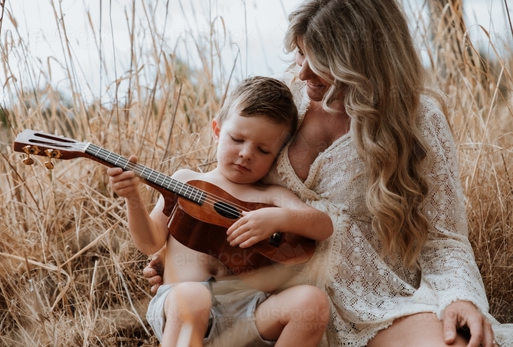 Mother and son sitting close together outside in field with small guitar - Australian Stock Image