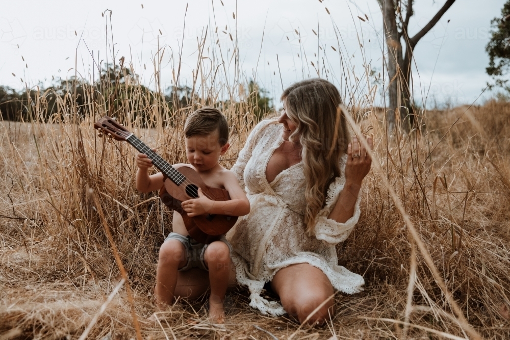 Mother and son sitting close together outside in field with small guitar - Australian Stock Image