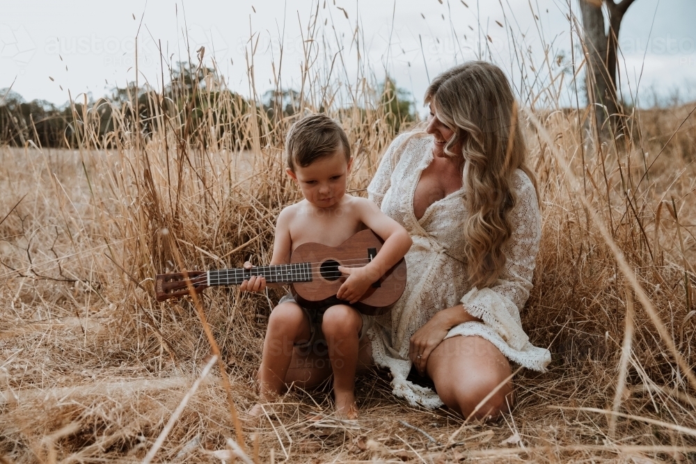 Mother and son sitting close together outside in field with small guitar - Australian Stock Image