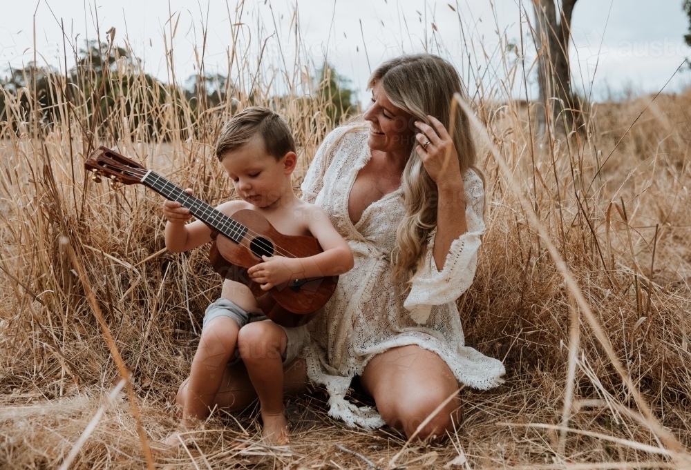 Mother and son sitting close together outside in field with small guitar - Australian Stock Image