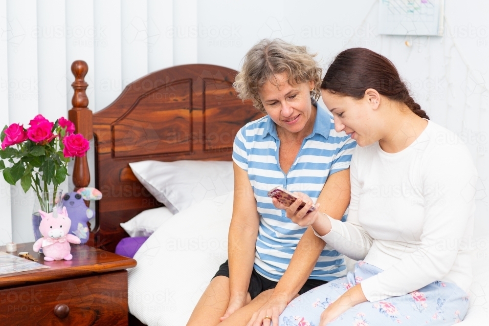 Mother and teen daughter sit-in non bed looking at smartphone - Australian Stock Image