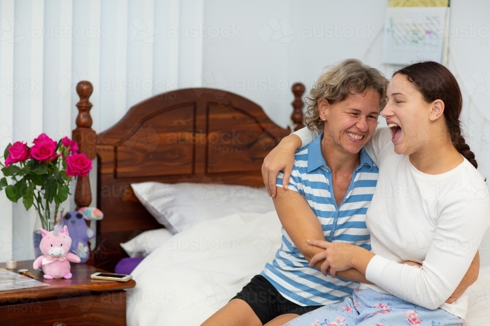 Mother and teen daughter sitting on bed and laughing with arms around each other - Australian Stock Image